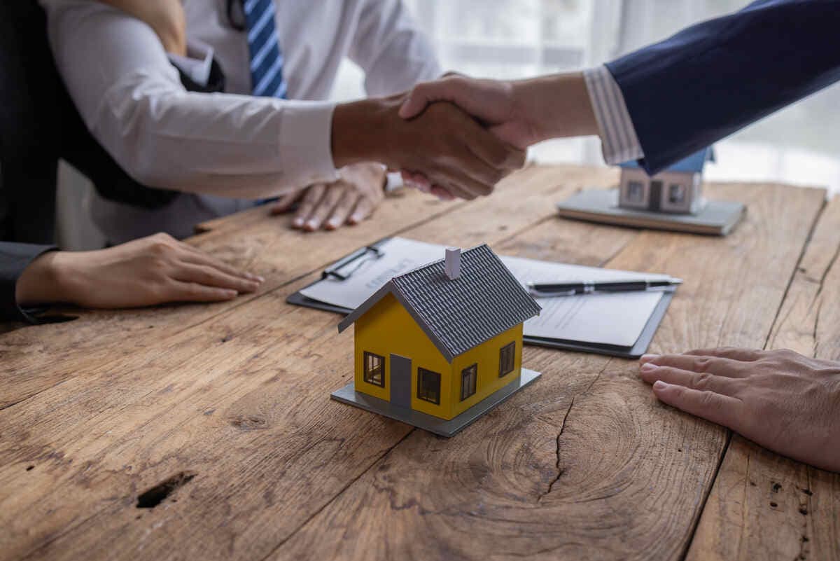 Yellow house model on table, handshake, and documents symbolizing a real estate deal.