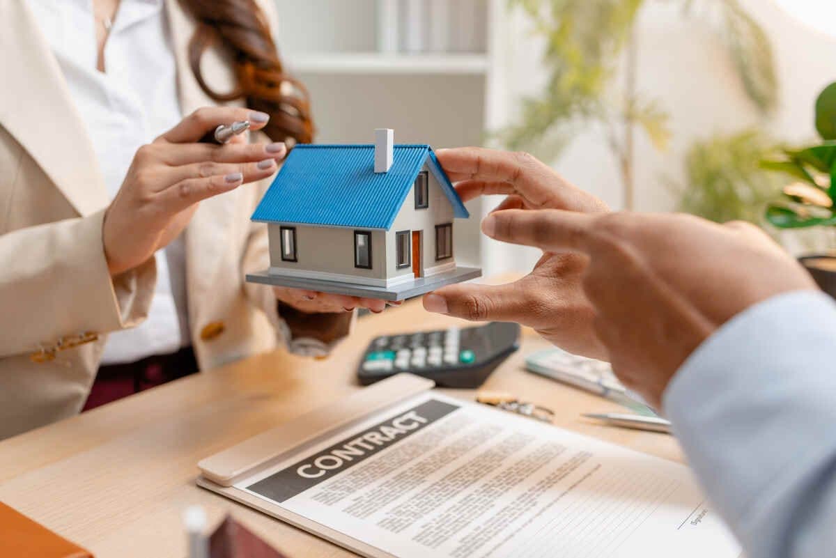 Two people exchange a small blue-roof house model over a contract on a desk.