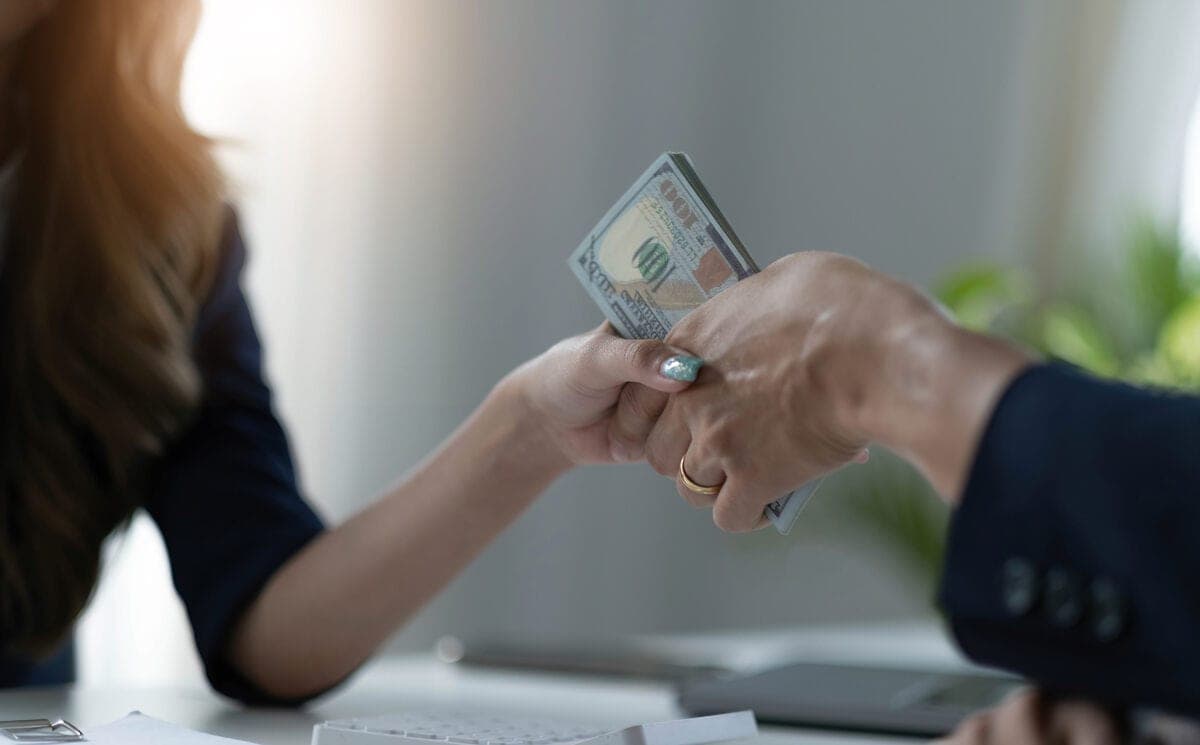 Close-up of two people exchanging US dollar bills in a bright office.