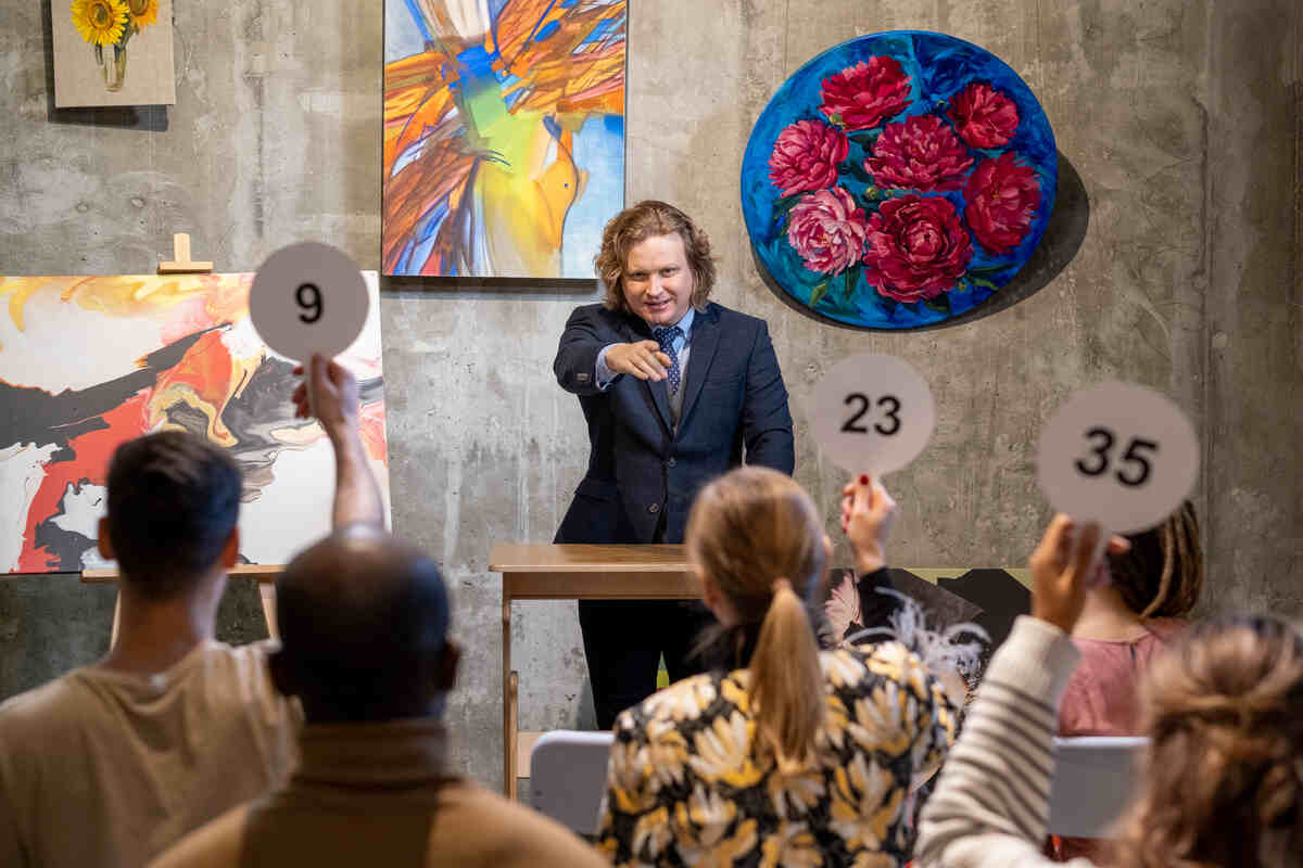 Auctioneer leads art sale as bidders raise paddles amid colorful paintings.