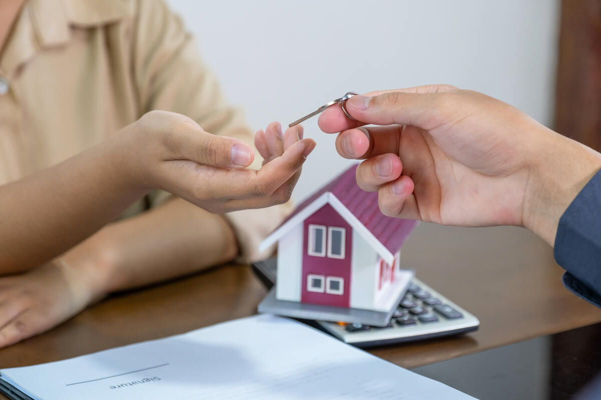 Hand passing a house key over paperwork beside a small house model.