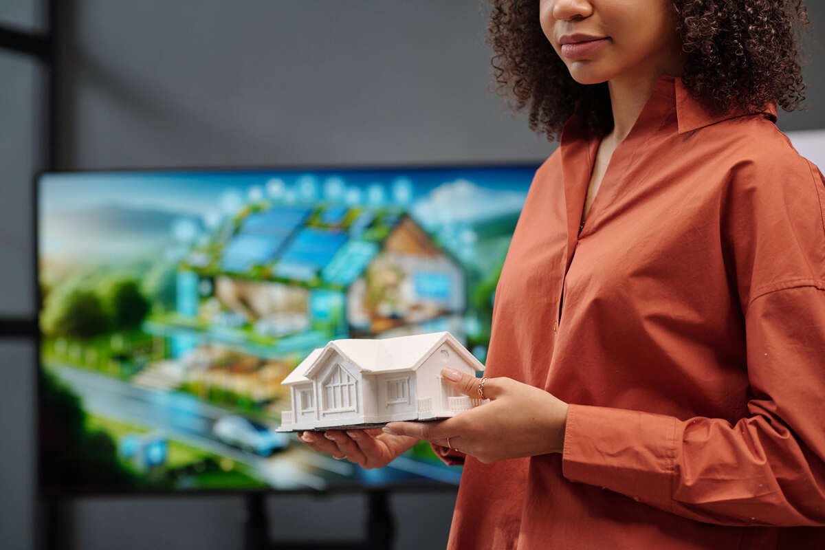 Person in a rust-colored shirt holds a house model before a blurred screen of a sustainable home.